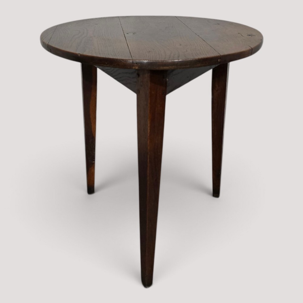 A dark brown Georgian oak cricket table featuring a circular planked top supported by three splayed tapered legs.