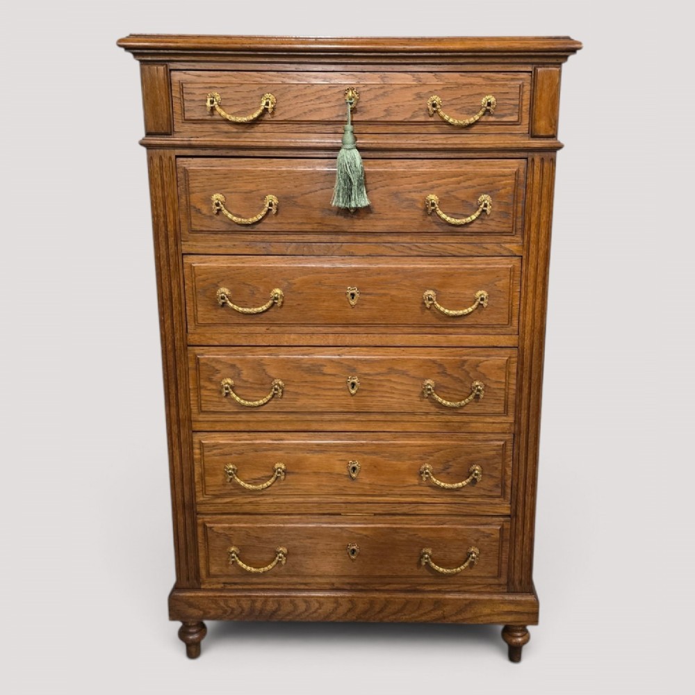 A tall French oak chest of drawers featuring six stacked drawers, brass swing handles, fluted side pilasters, and turned wooden feet.