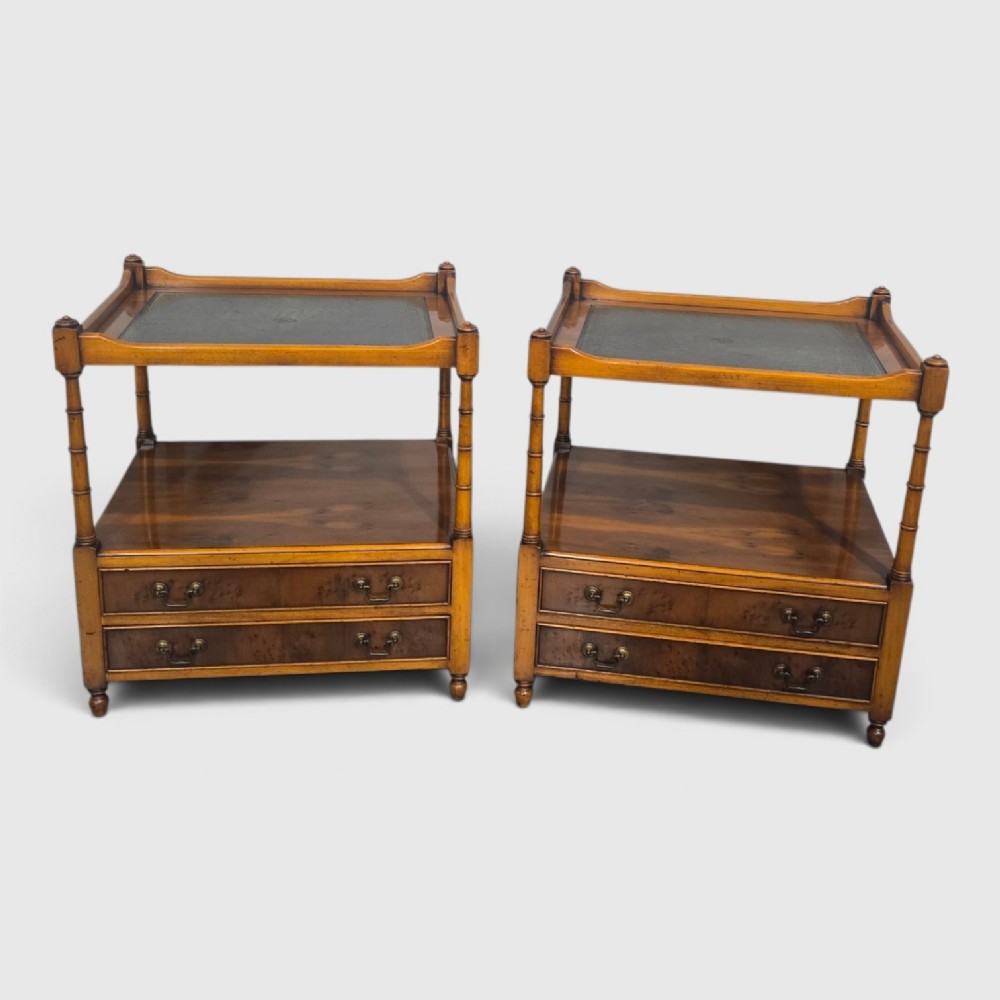 A pair of yew wood lamp tables featuring dark green leather inset tops, a middle shelf, two drawers with brass handles, and turned corner columns.