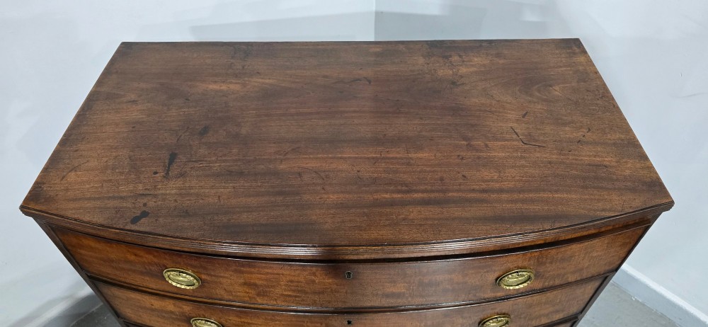 A Georgian mahogany bowfront chest of drawers featuring four graduated drawers with oval brass handles and splayed feet. View IX