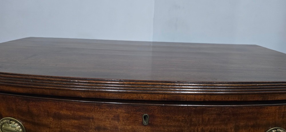 A Georgian mahogany bowfront chest of drawers featuring four graduated drawers with oval brass handles and splayed feet. View X