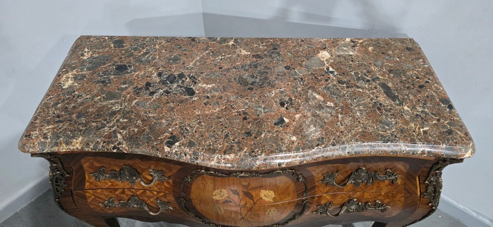 A French Louis XV style bombe commode featuring detailed floral marquetry inlay, bronze mounts, curved legs, and a brown marble top. View IX