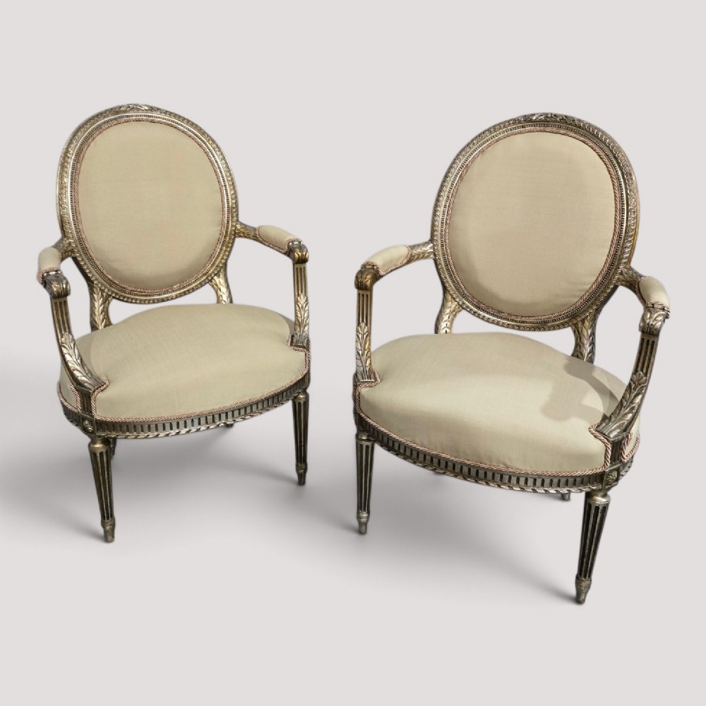 A pair of French Louis XVI style open armchairs featuring carved giltwood frames with oval medallion backs, fluted legs, and neutral cream upholstery.