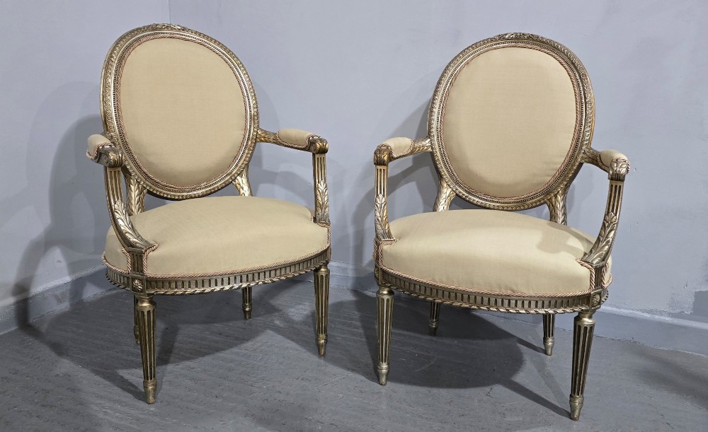 A pair of French Louis XVI style open armchairs featuring carved giltwood frames with oval medallion backs, fluted legs, and neutral cream upholstery. View II