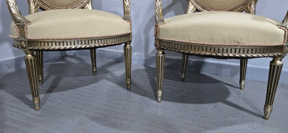 A pair of French Louis XVI style open armchairs featuring carved giltwood frames with oval medallion backs, fluted legs, and neutral cream upholstery. View III