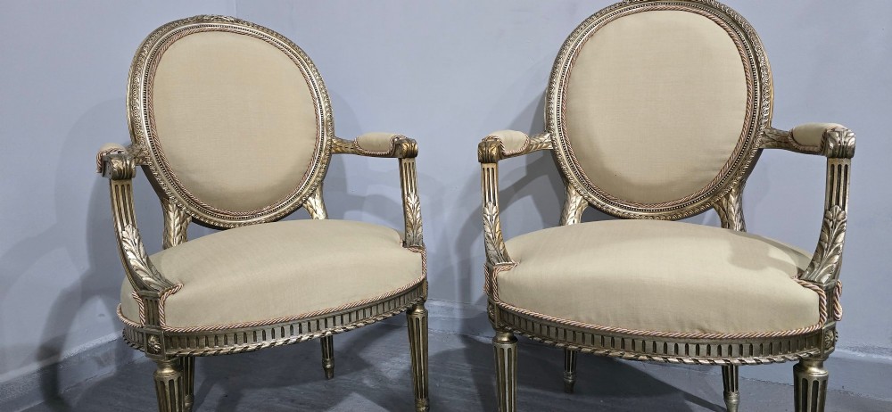 A pair of French Louis XVI style open armchairs featuring carved giltwood frames with oval medallion backs, fluted legs, and neutral cream upholstery. View IV
