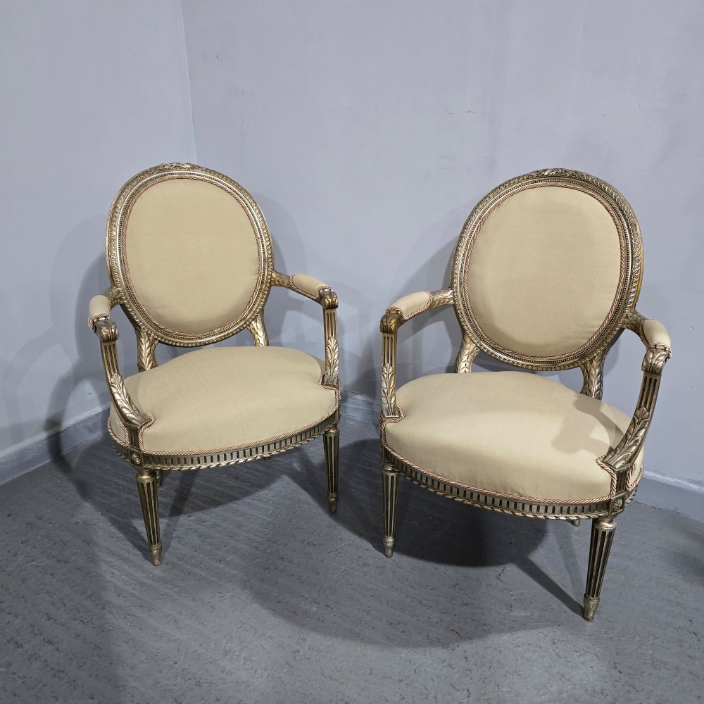 A pair of French Louis XVI style open armchairs featuring carved giltwood frames with oval medallion backs, fluted legs, and neutral cream upholstery. View VI
