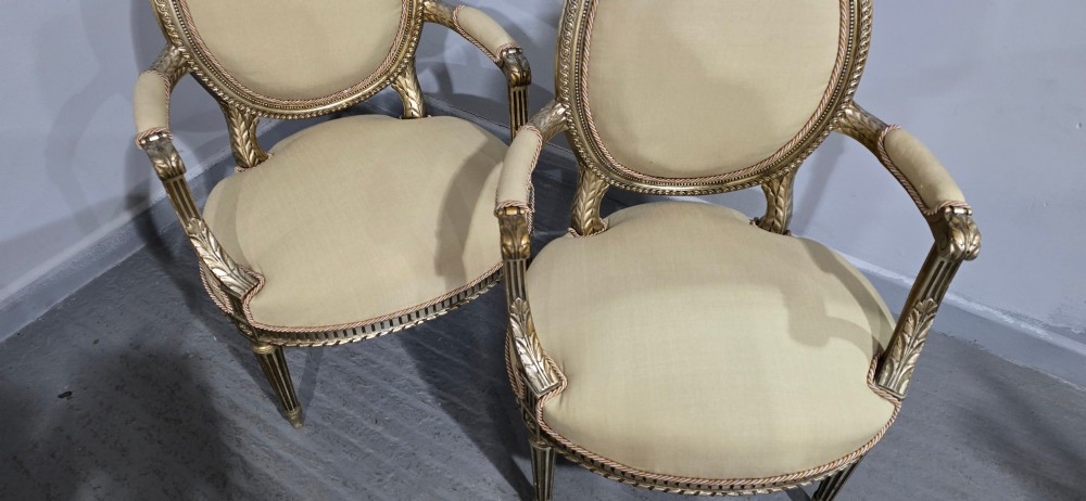 A pair of French Louis XVI style open armchairs featuring carved giltwood frames with oval medallion backs, fluted legs, and neutral cream upholstery. View XI