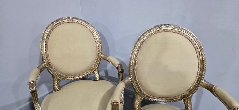 A pair of French Louis XVI style open armchairs featuring carved giltwood frames with oval medallion backs, fluted legs, and neutral cream upholstery. View XII