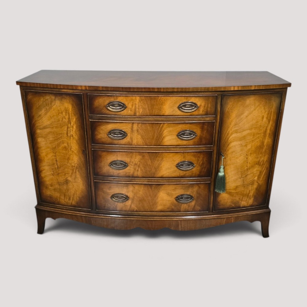 A bow-front mahogany sideboard featuring four central drawers flanked by two side cupboard doors with oval brass handles and flame mahogany grain patterns.