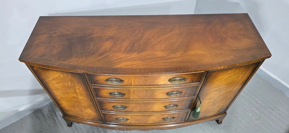 A bow-front mahogany sideboard featuring four central drawers flanked by two side cupboard doors with oval brass handles and flame mahogany grain patterns. View IV