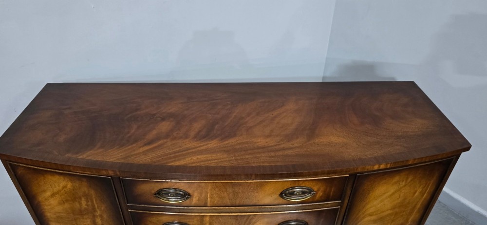 A bow-front mahogany sideboard featuring four central drawers flanked by two side cupboard doors with oval brass handles and flame mahogany grain patterns. View V