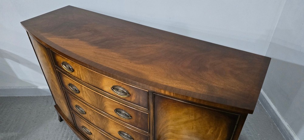 A bow-front mahogany sideboard featuring four central drawers flanked by two side cupboard doors with oval brass handles and flame mahogany grain patterns. View VI
