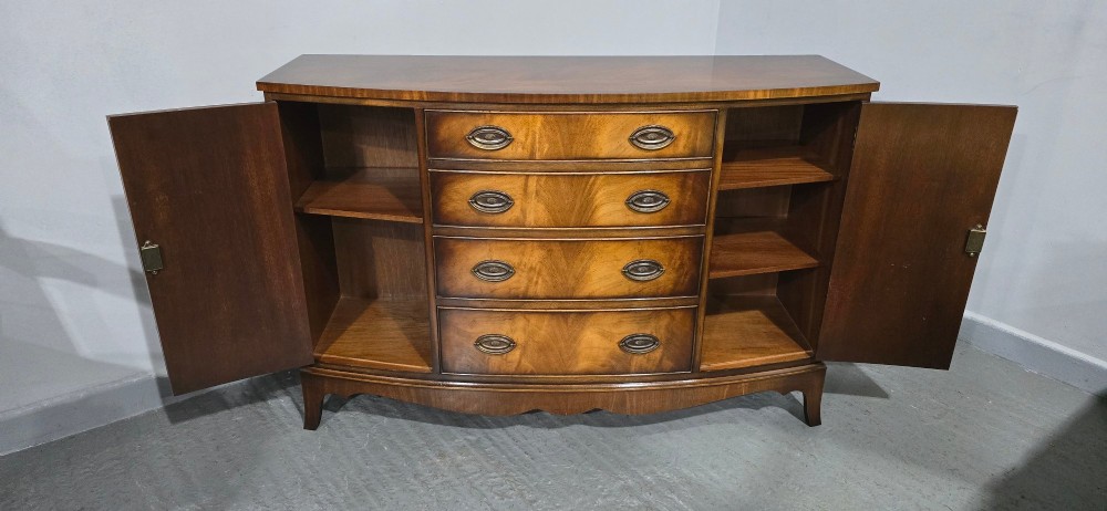 A bow-front mahogany sideboard featuring four central drawers flanked by two side cupboard doors with oval brass handles and flame mahogany grain patterns. View X