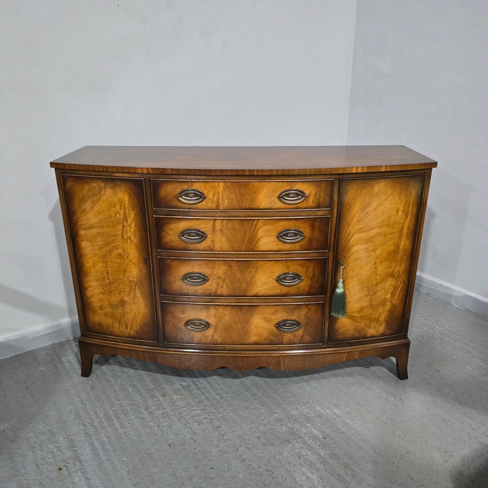 A bow-front mahogany sideboard featuring four central drawers flanked by two side cupboard doors with oval brass handles and flame mahogany grain patterns. View XIII