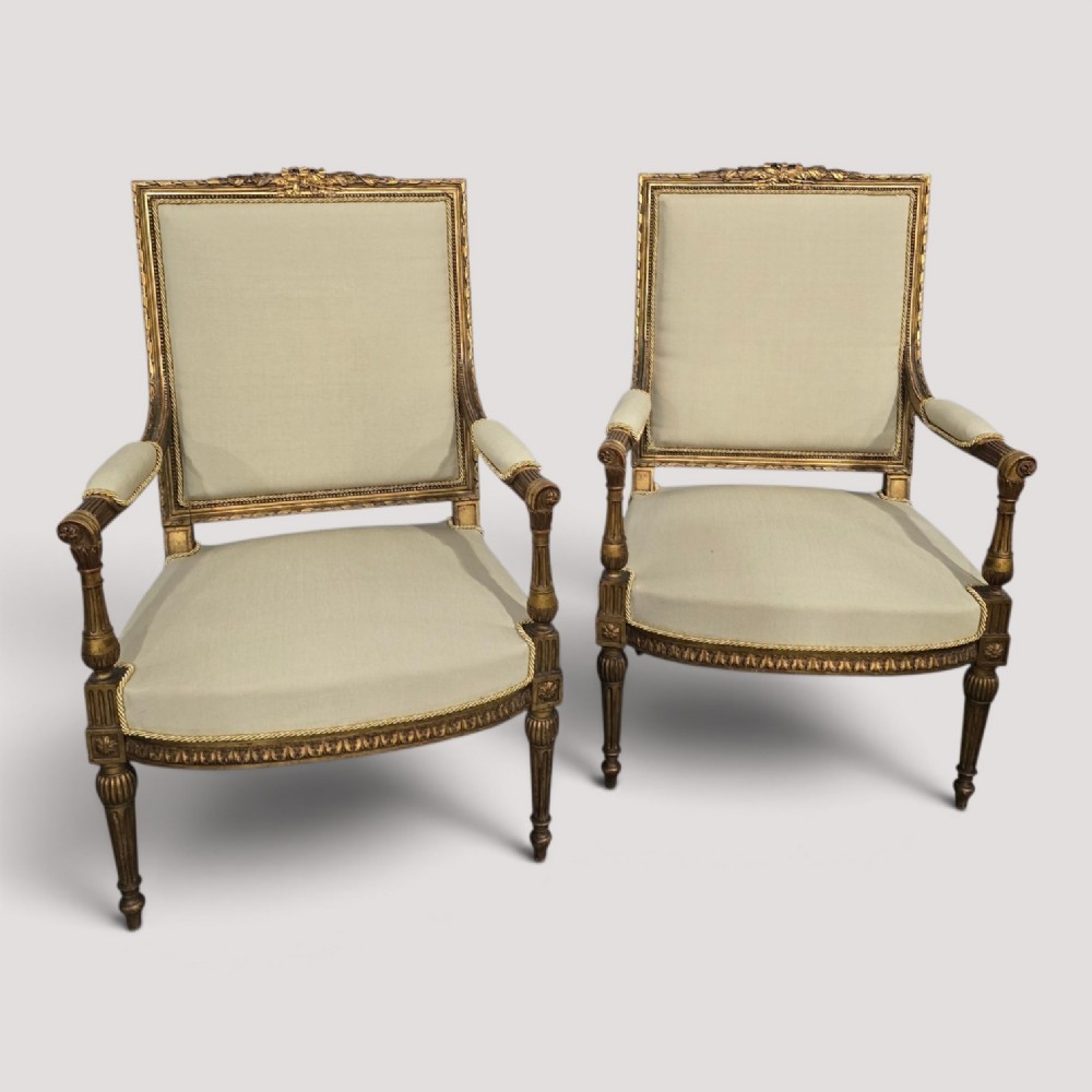 A pair of French Neoclassical giltwood open armchairs upholstered in neutral fabric with fluted tapered legs and carved square backs.