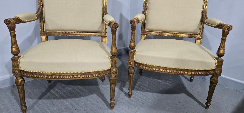 A pair of French Neoclassical giltwood open armchairs upholstered in neutral fabric with fluted tapered legs and carved square backs. View IV