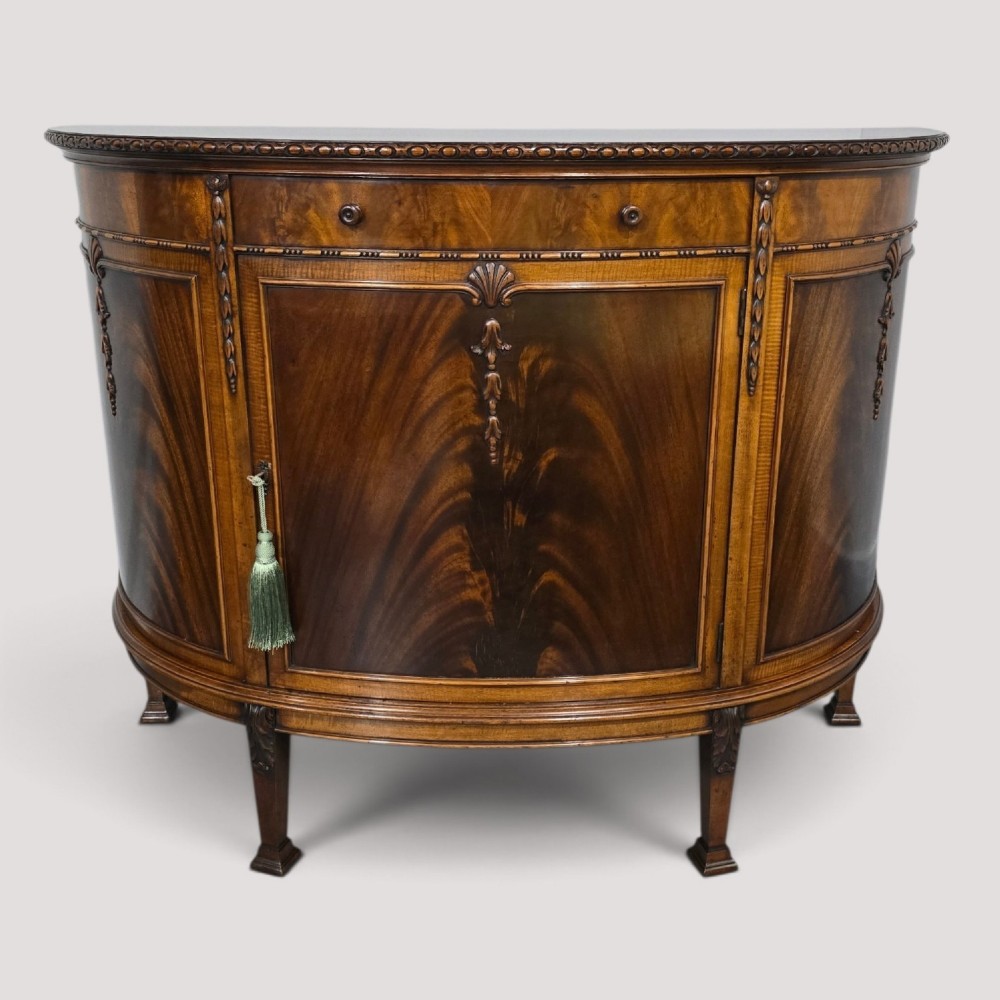 A semi-circular mahogany demi-lune side cabinet featuring flame mahogany door panels, carved details, a single upper drawer, and tapered square feet.