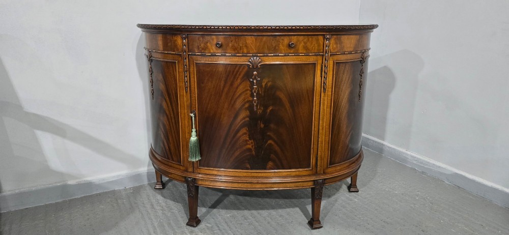 A semi-circular mahogany demi-lune side cabinet featuring flame mahogany door panels, carved details, a single upper drawer, and tapered square feet. View II