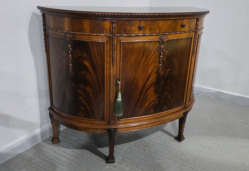 A semi-circular mahogany demi-lune side cabinet featuring flame mahogany door panels, carved details, a single upper drawer, and tapered square feet. View III