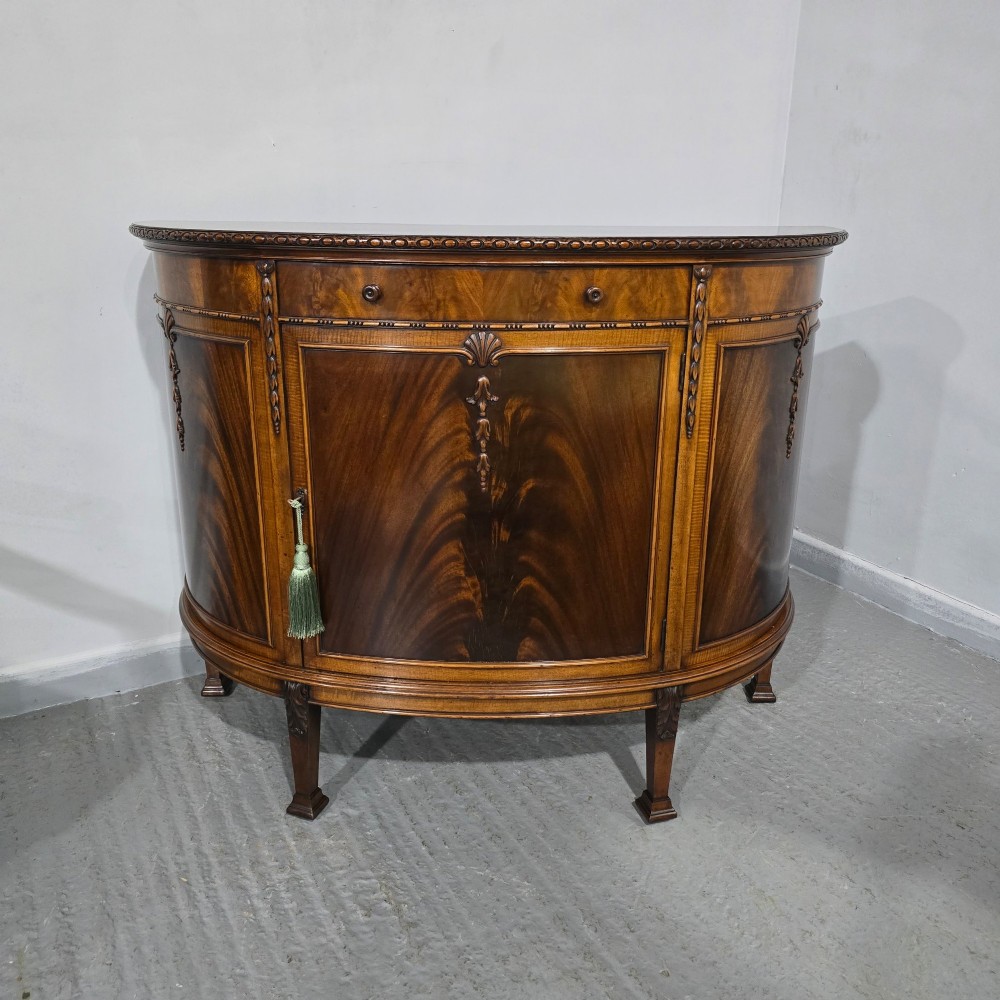 A semi-circular mahogany demi-lune side cabinet featuring flame mahogany door panels, carved details, a single upper drawer, and tapered square feet. View VI