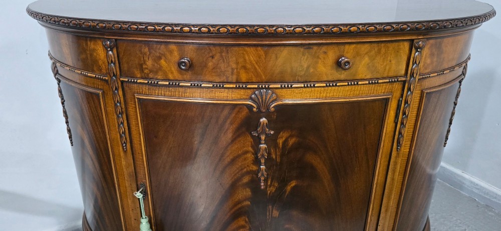 A semi-circular mahogany demi-lune side cabinet featuring flame mahogany door panels, carved details, a single upper drawer, and tapered square feet. View VII