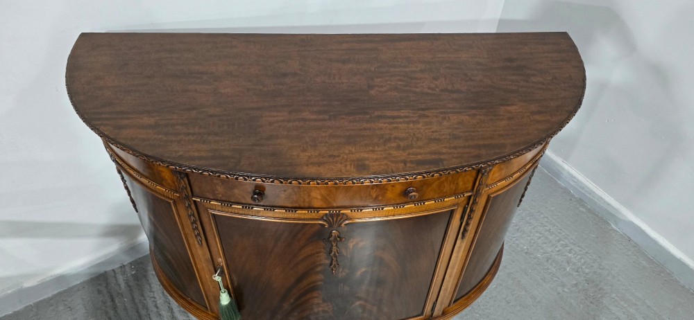 A semi-circular mahogany demi-lune side cabinet featuring flame mahogany door panels, carved details, a single upper drawer, and tapered square feet. View IX