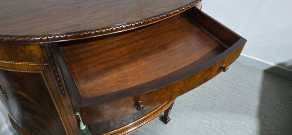 A semi-circular mahogany demi-lune side cabinet featuring flame mahogany door panels, carved details, a single upper drawer, and tapered square feet. View X