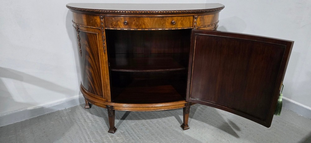 A semi-circular mahogany demi-lune side cabinet featuring flame mahogany door panels, carved details, a single upper drawer, and tapered square feet. View XI