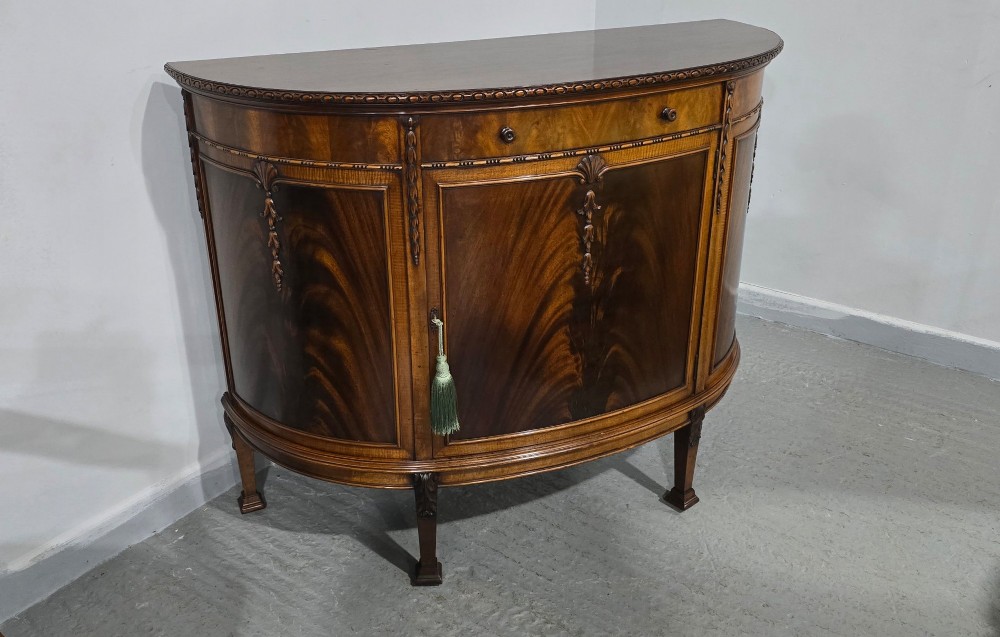 A semi-circular mahogany demi-lune side cabinet featuring flame mahogany door panels, carved details, a single upper drawer, and tapered square feet. View XII
