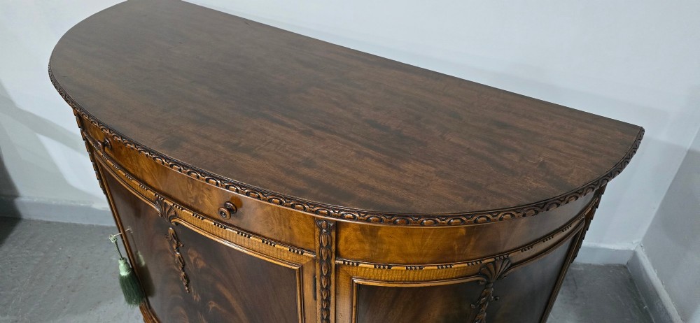 A semi-circular mahogany demi-lune side cabinet featuring flame mahogany door panels, carved details, a single upper drawer, and tapered square feet. View XIV
