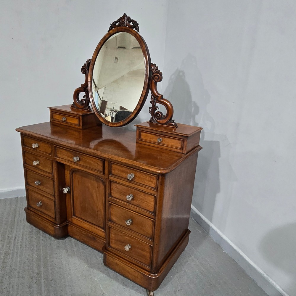 A Victorian mahogany dressing table featuring a central oval swivelling mirror, carved scroll supports, small upper trinket drawers, and twin drawer pedestals flanking a central cupboard. View XII