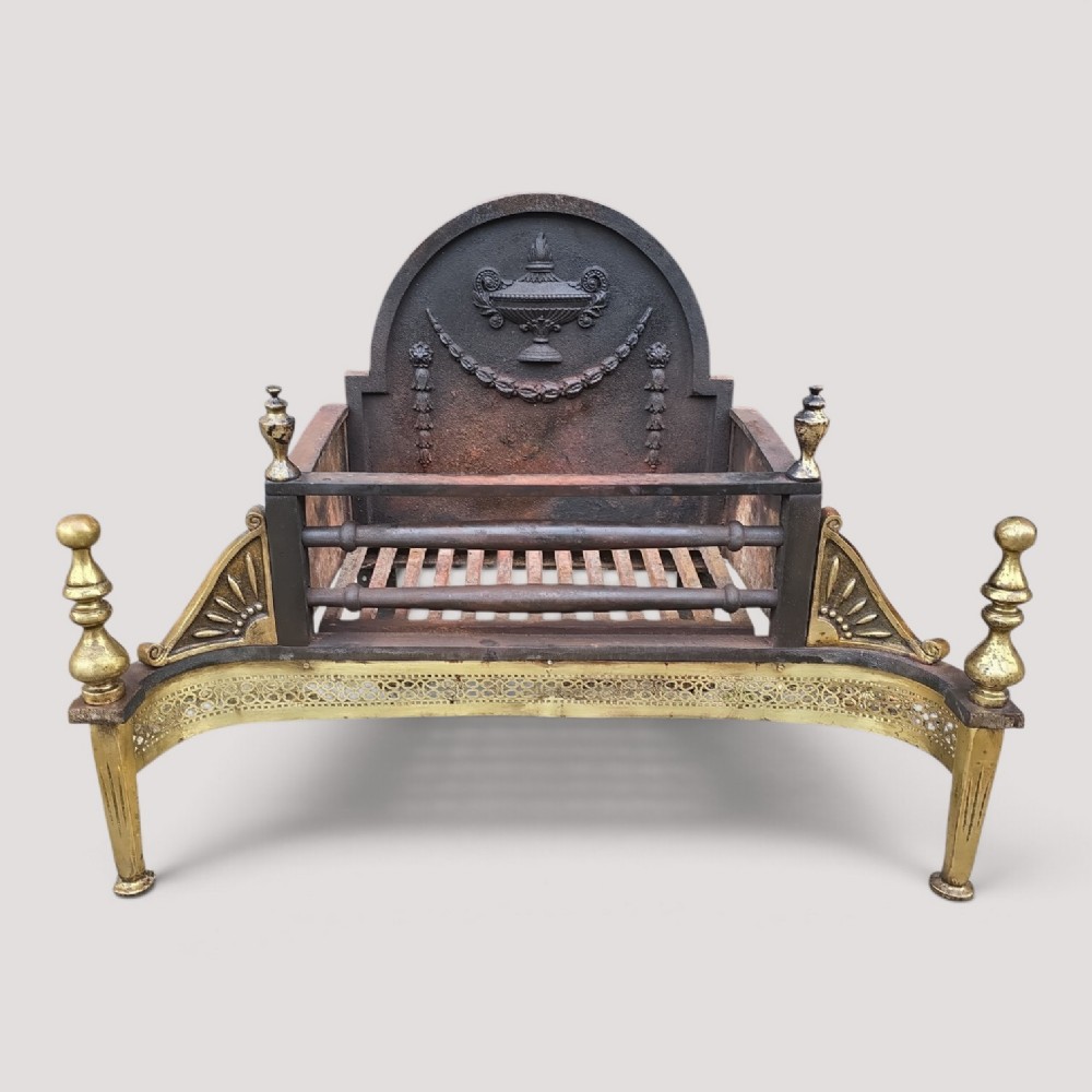An Adam style fireplace dog grate crafted from cast iron and brass, featuring a serpentine pierced apron, urn finials, and a relief urn backplate.
