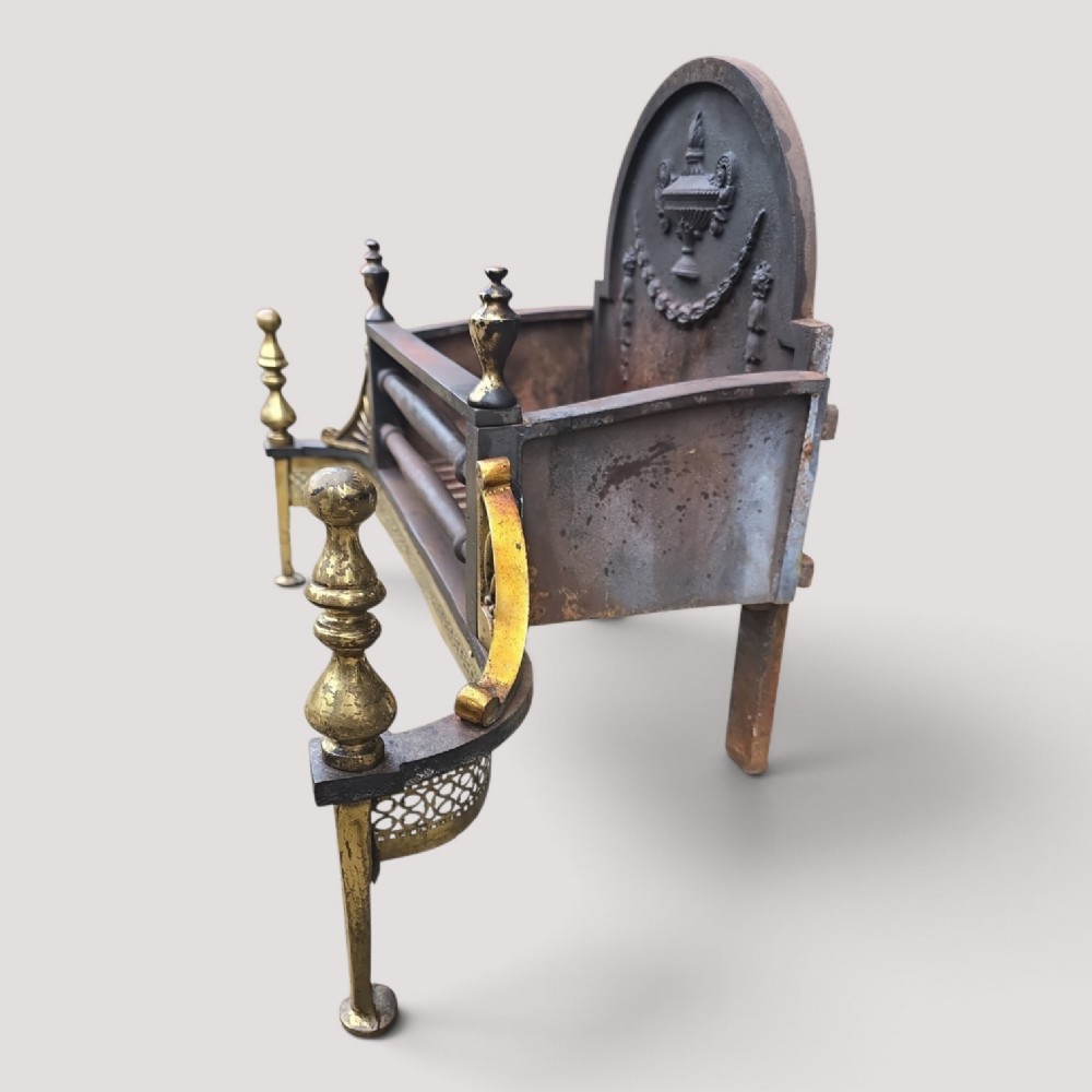 An Adam style fireplace dog grate crafted from cast iron and brass, featuring a serpentine pierced apron, urn finials, and a relief urn backplate. View III