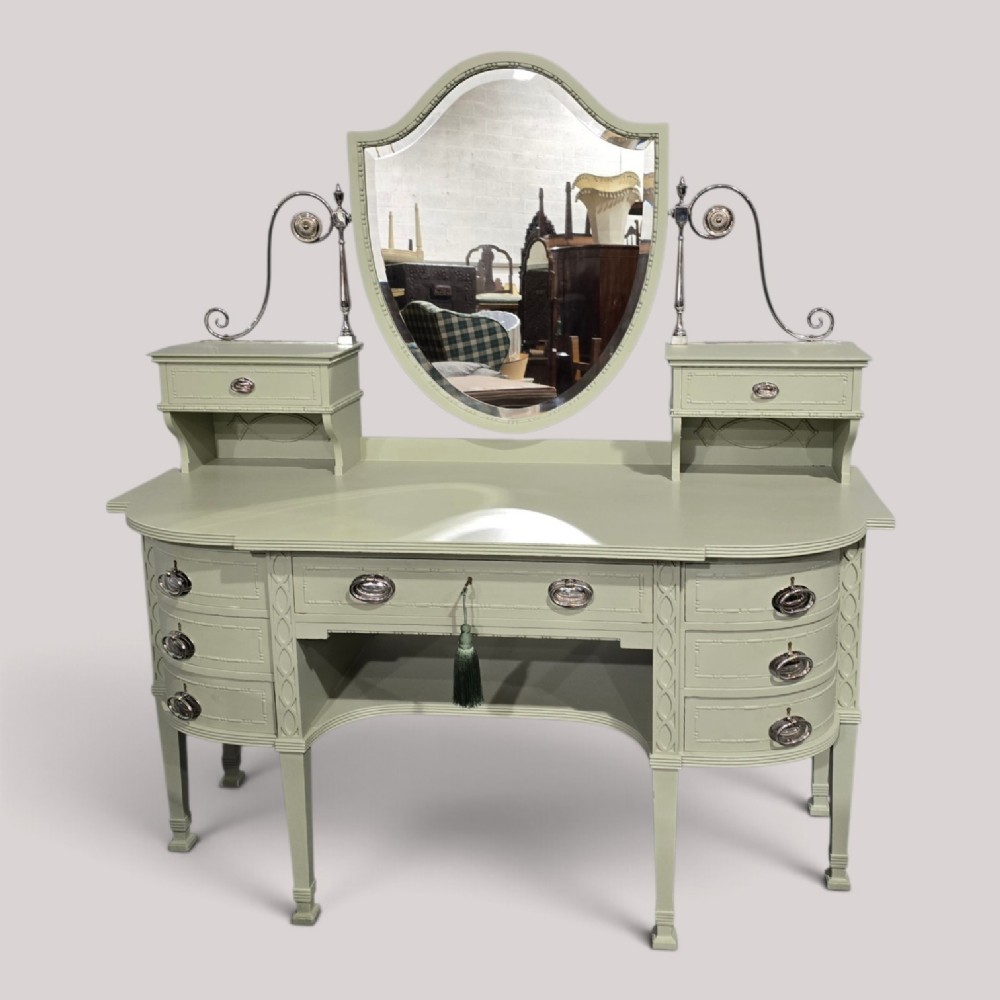 A sage green painted dressing table featuring an integrated central shield mirror, side trinket drawers, metal scroll sconces, and tapered legs.
