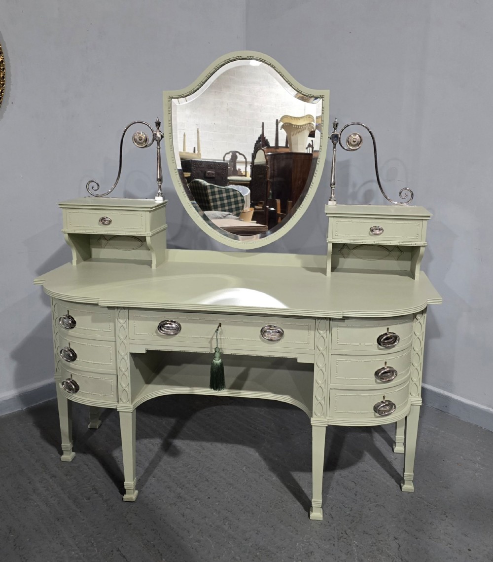 A sage green painted dressing table featuring an integrated central shield mirror, side trinket drawers, metal scroll sconces, and tapered legs. View II