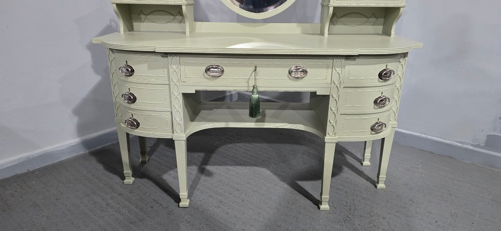 A sage green painted dressing table featuring an integrated central shield mirror, side trinket drawers, metal scroll sconces, and tapered legs. View III
