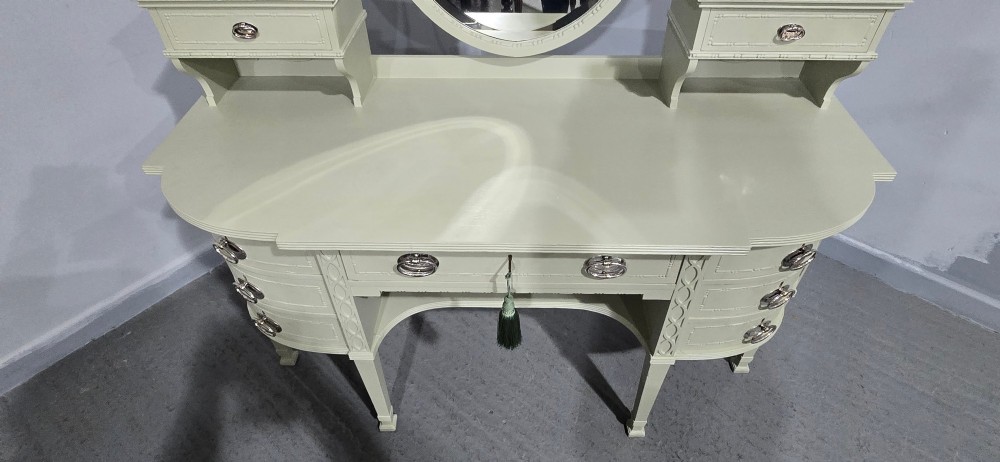 A sage green painted dressing table featuring an integrated central shield mirror, side trinket drawers, metal scroll sconces, and tapered legs. View XI