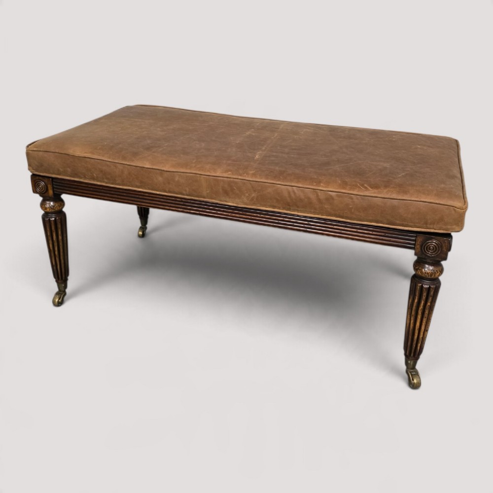 A rectangular Regency bench featuring tan leather upholstery, a reeded dark wood frame, tapered carved legs, and brass castors.