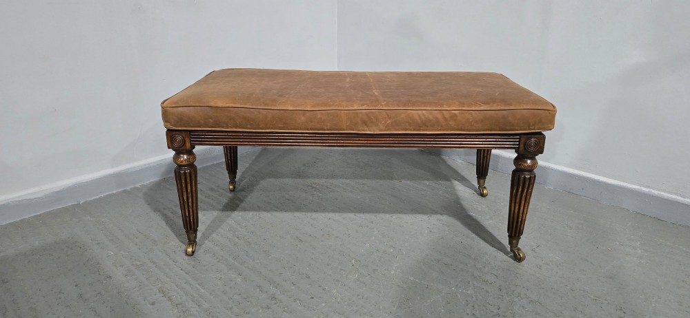 A rectangular Regency bench featuring tan leather upholstery, a reeded dark wood frame, tapered carved legs, and brass castors. View II