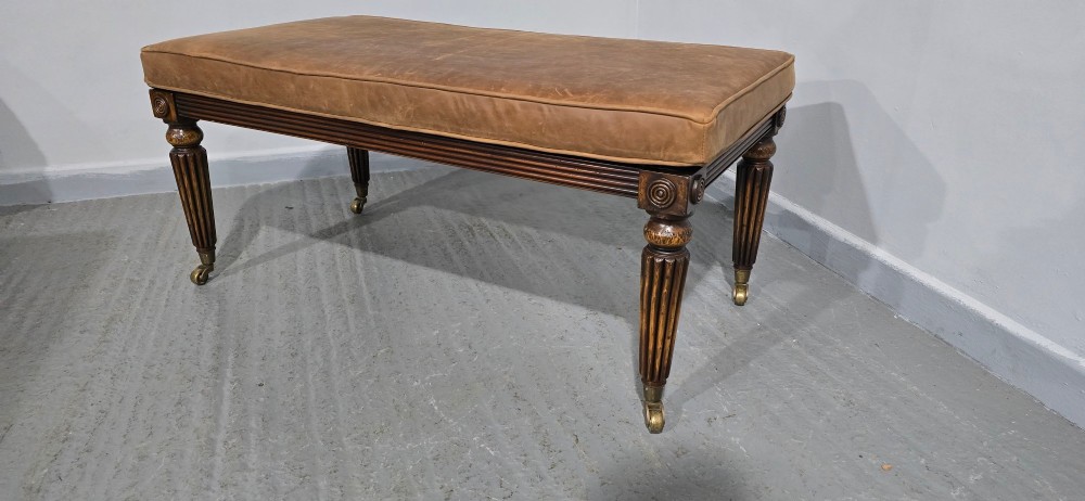 A rectangular Regency bench featuring tan leather upholstery, a reeded dark wood frame, tapered carved legs, and brass castors. View IV