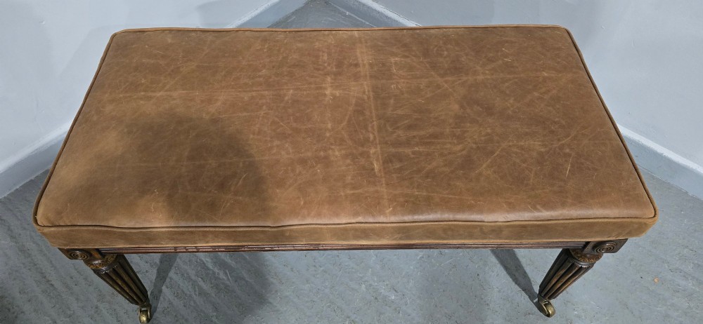 A rectangular Regency bench featuring tan leather upholstery, a reeded dark wood frame, tapered carved legs, and brass castors. View V