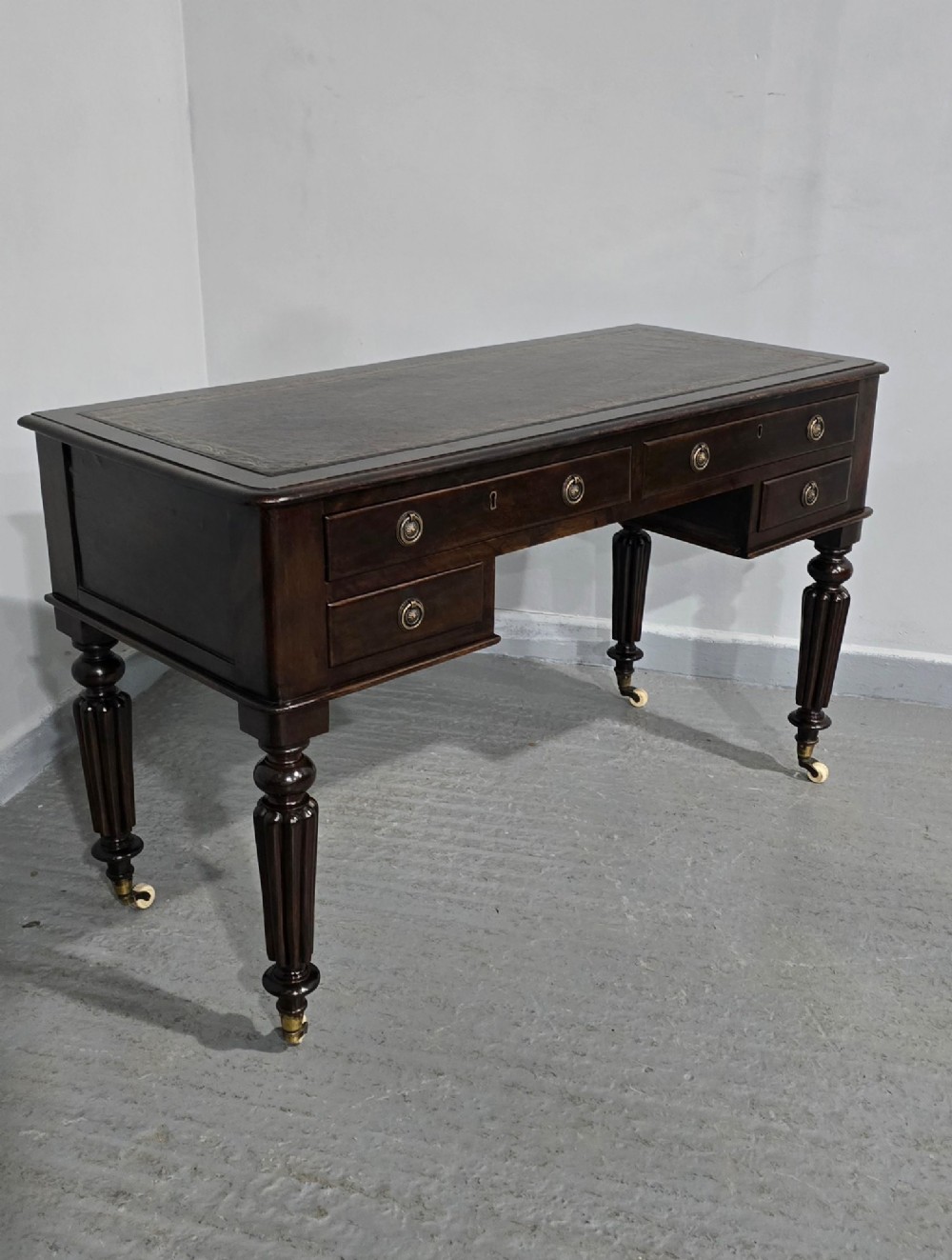 A Victorian dark mahogany writing desk featuring a green leather inset top, four drawers with circular brass ring handles, turned fluted legs, and brass castors. View VIII