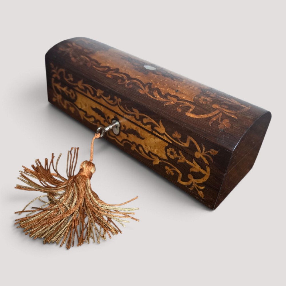An antique dark wood rectangular glove box featuring foliate marquetry inlay, a curved lid, a key with a silk tassel, and a mother-of-pearl escutcheon.