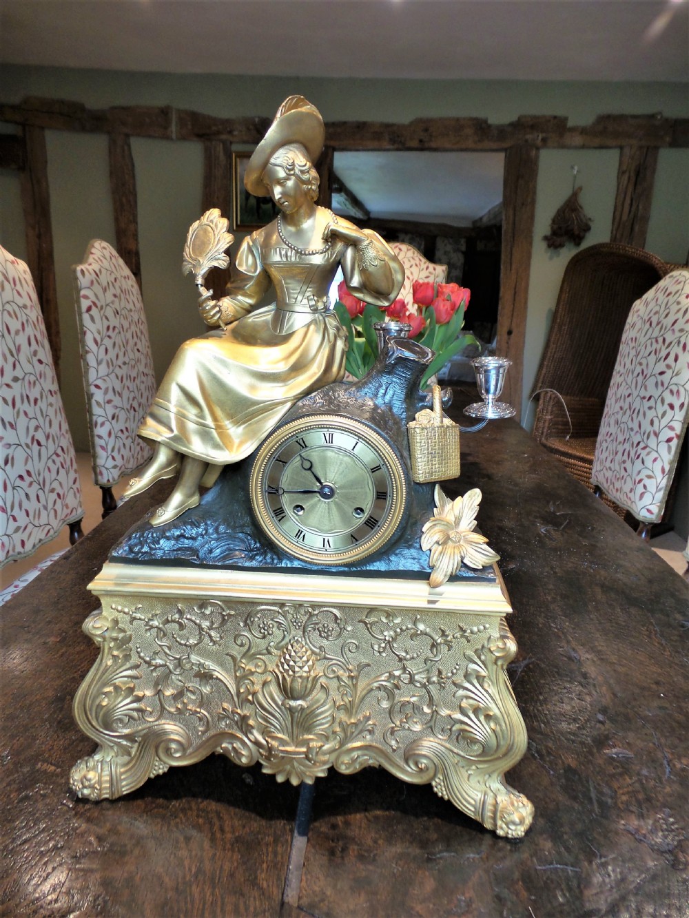 A mid-19th-century French gilt and patinated bronze mantel clock featuring a sculpted female figure holding a fan and seated on a tree stump atop an ornate scroll-carved base.