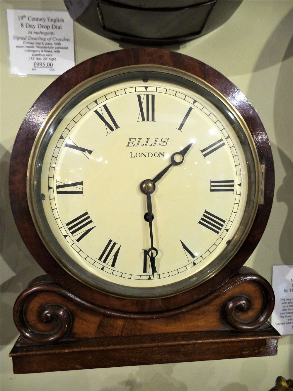 A 19th-century mahogany dial clock featuring a white circular face signed Ellis London with Roman numerals, a brass bezel, and carved scroll detailing at the base.