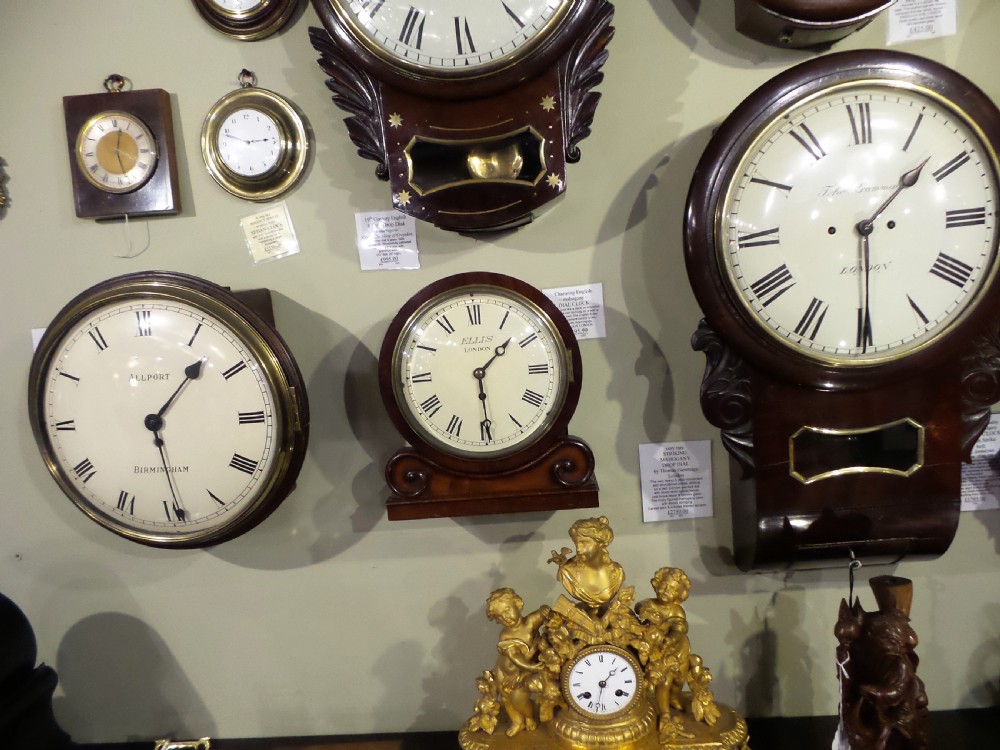 A 19th-century mahogany dial clock featuring a white circular face signed Ellis London with Roman numerals, a brass bezel, and carved scroll detailing at the base. View II