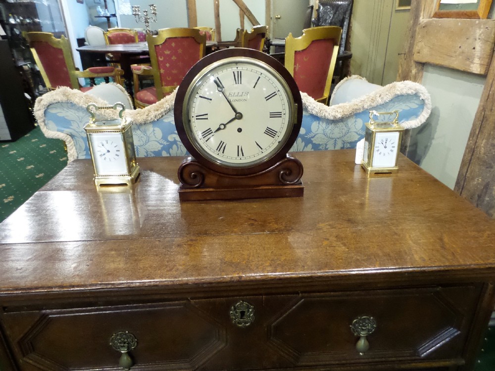 A 19th-century mahogany dial clock featuring a white circular face signed Ellis London with Roman numerals, a brass bezel, and carved scroll detailing at the base. View V