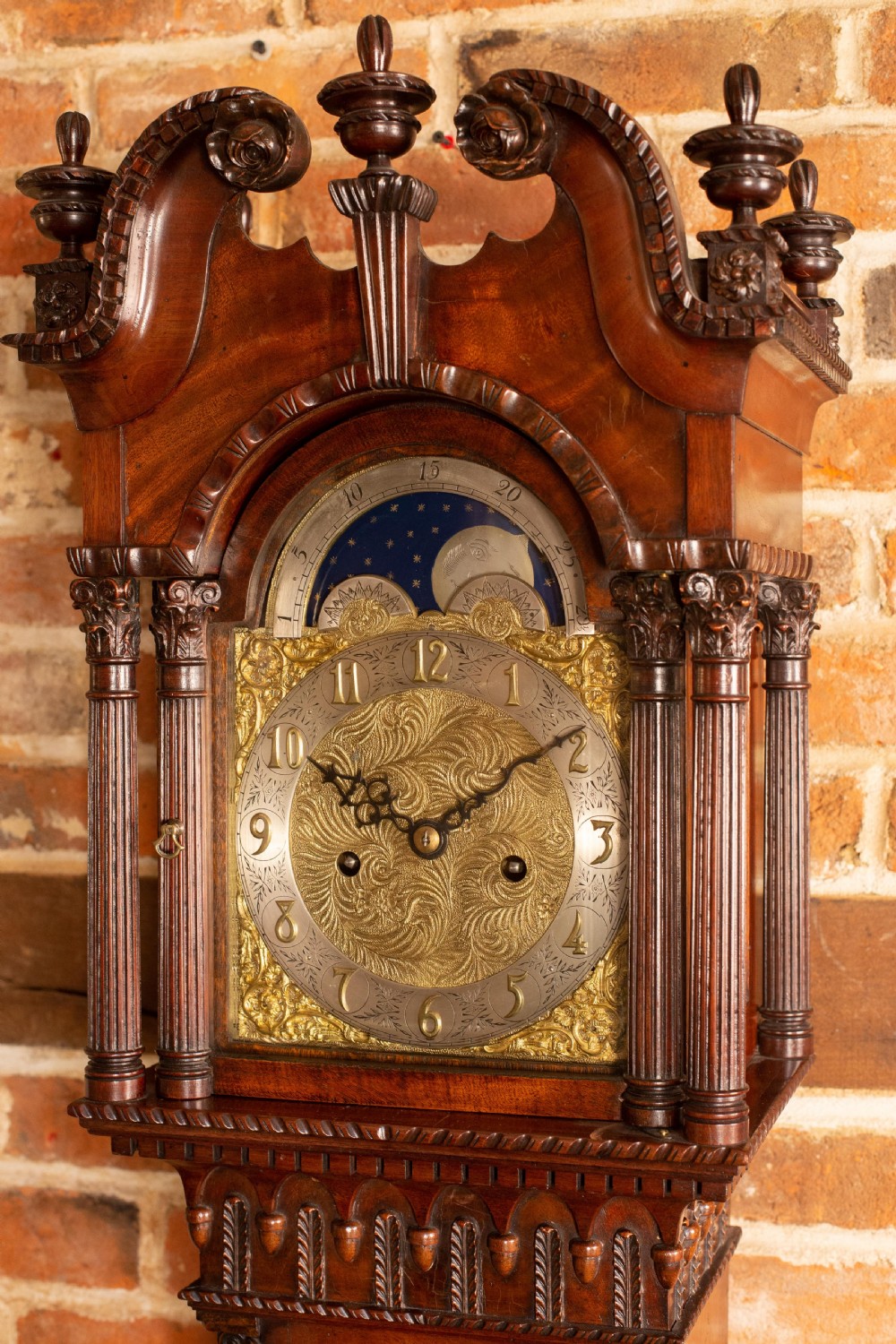 An Edwardian miniature mahogany longcase clock featuring a carved swan-neck pediment, brass moon phase dial, and glass trunk door revealing brass weights. View III