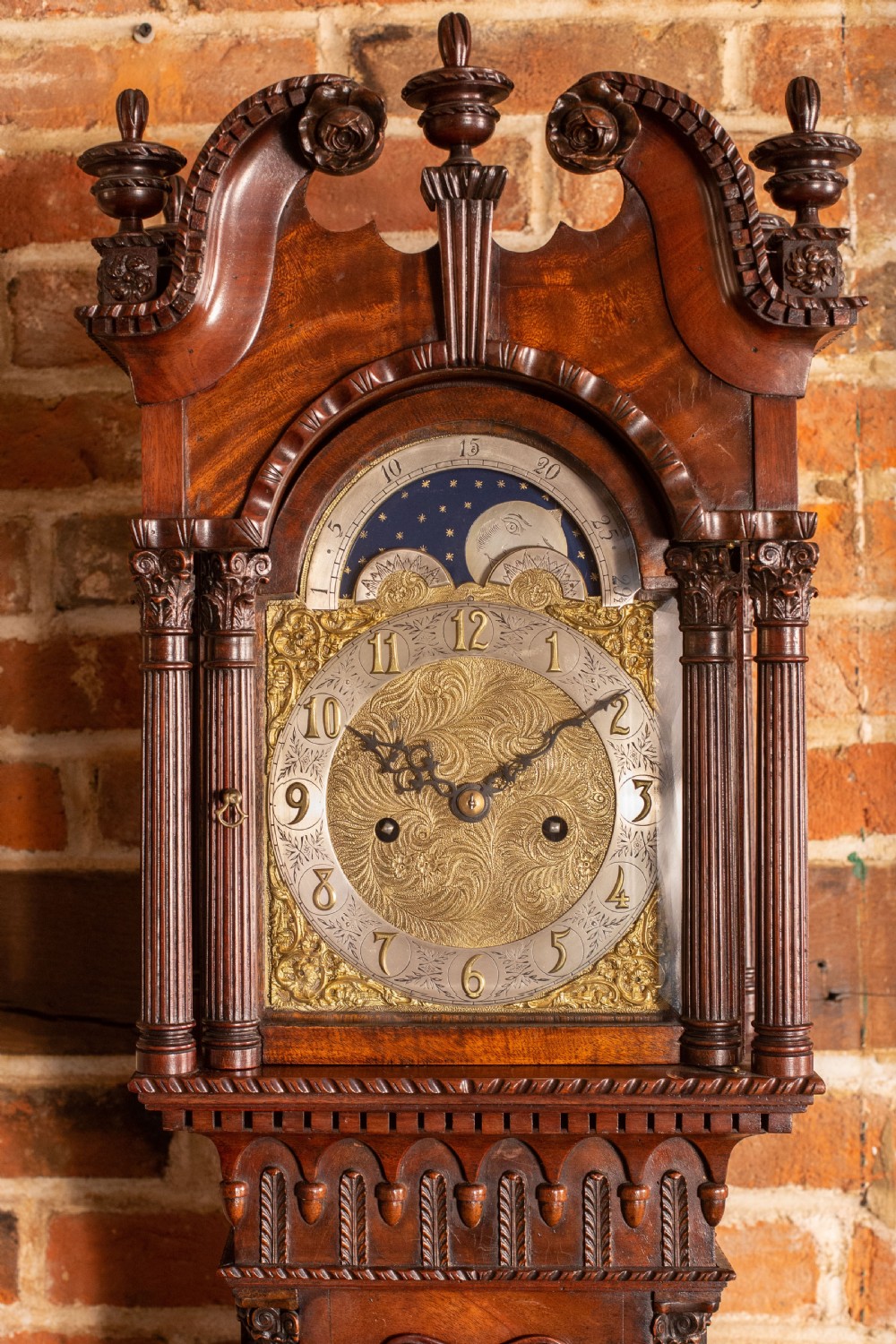 An Edwardian miniature mahogany longcase clock featuring a carved swan-neck pediment, brass moon phase dial, and glass trunk door revealing brass weights. View IV